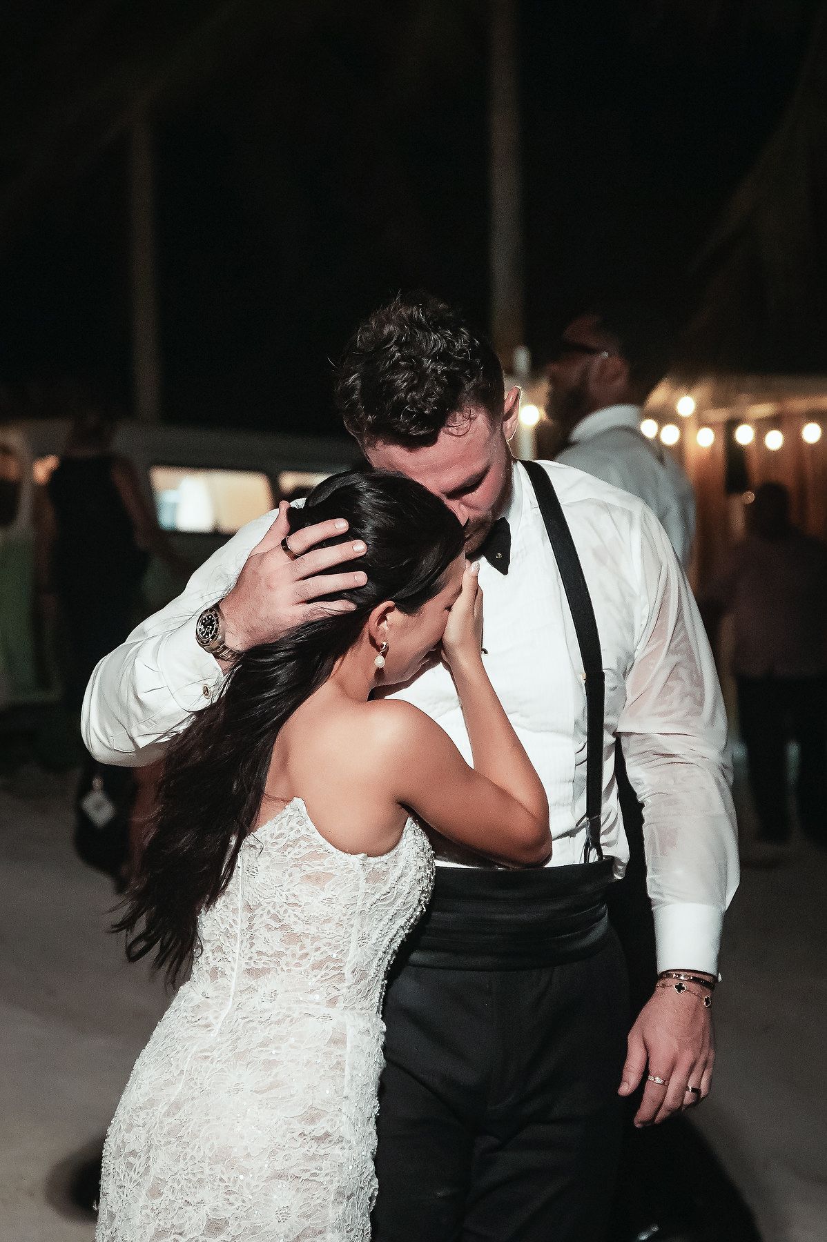 Bride and groom share an emotional first dance embrace at outdoor wedding reception with string lights at night.