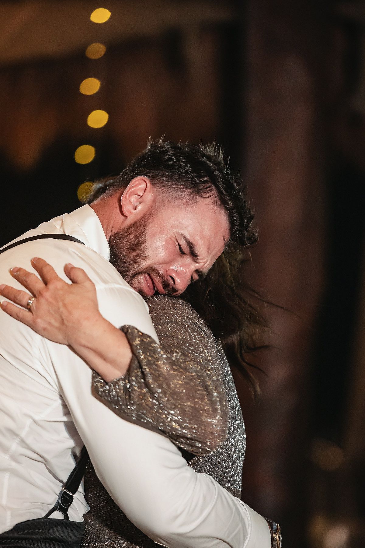 Emotional groom hugging a guest in a sparkly dress at a wedding reception with bokeh lights in background.
