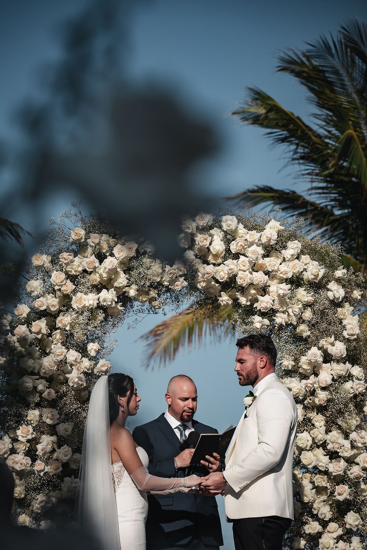 Couple exchanges vows under a floral arch of white roses with a officiant at an outdoor wedding ceremony.