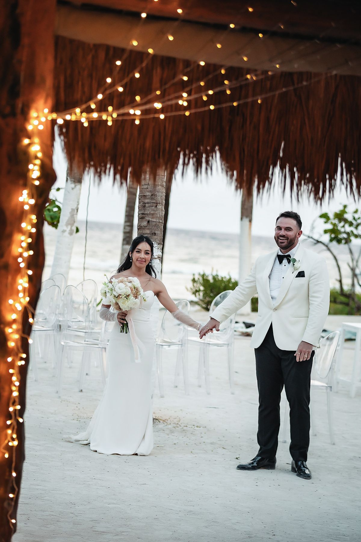 Bride and groom hold hands at a romantic beach wedding ceremony under a palapa adorned with string lights at dusk.