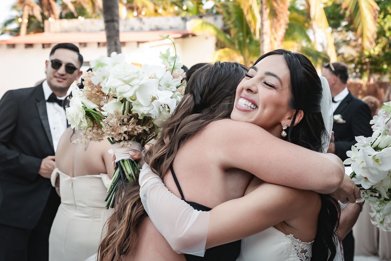 Bride hugging a guest at an outdoor wedding reception, holding a white bouquet, with guests in formal attire nearby.