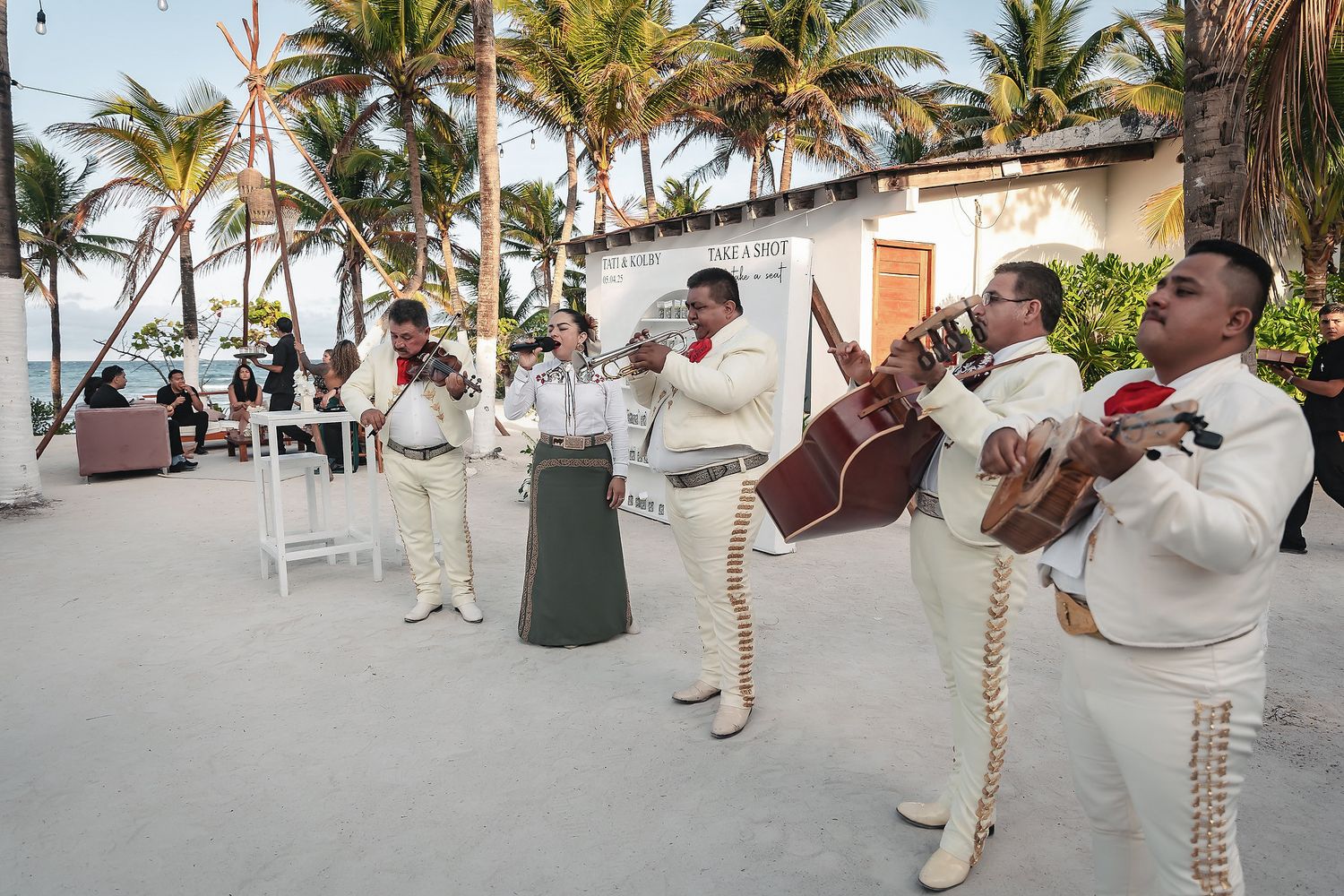 Mariachi band in traditional charro suits performing outdoors near a tropical beach with palm trees.