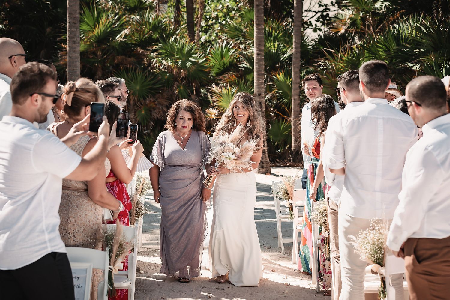 Bride walks down outdoor aisle with mother at tropical wedding ceremony, guests photographing the moment.