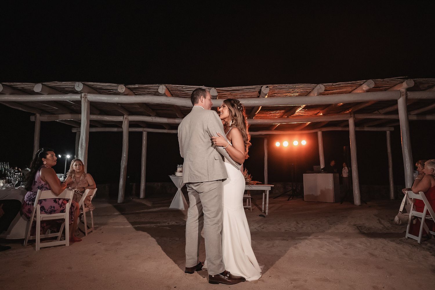 Couple sharing first dance at outdoor beach wedding reception under pergola at night with guests seated nearby.