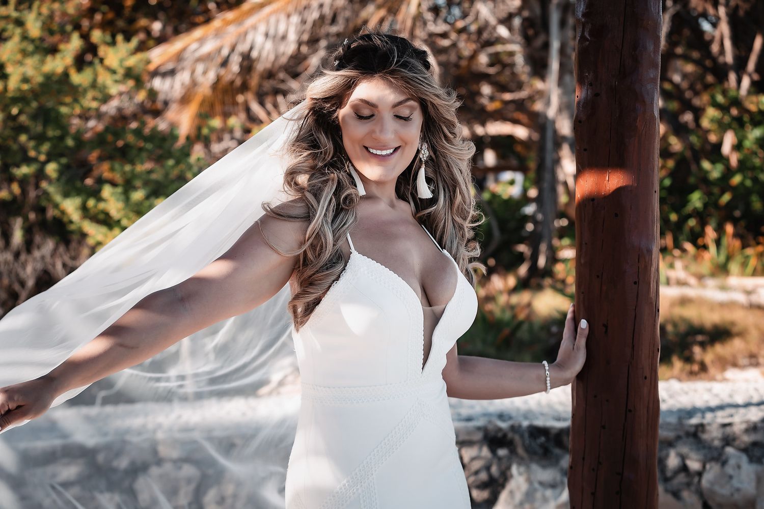 Smiling bride in white gown and flowing veil twirling outdoors near a wooden post on a sunny day.