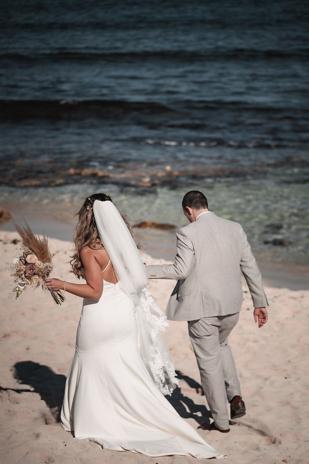 Bride and groom walking hand in hand on a sandy beach, bride holding a boho bouquet with veil flowing in the breeze.