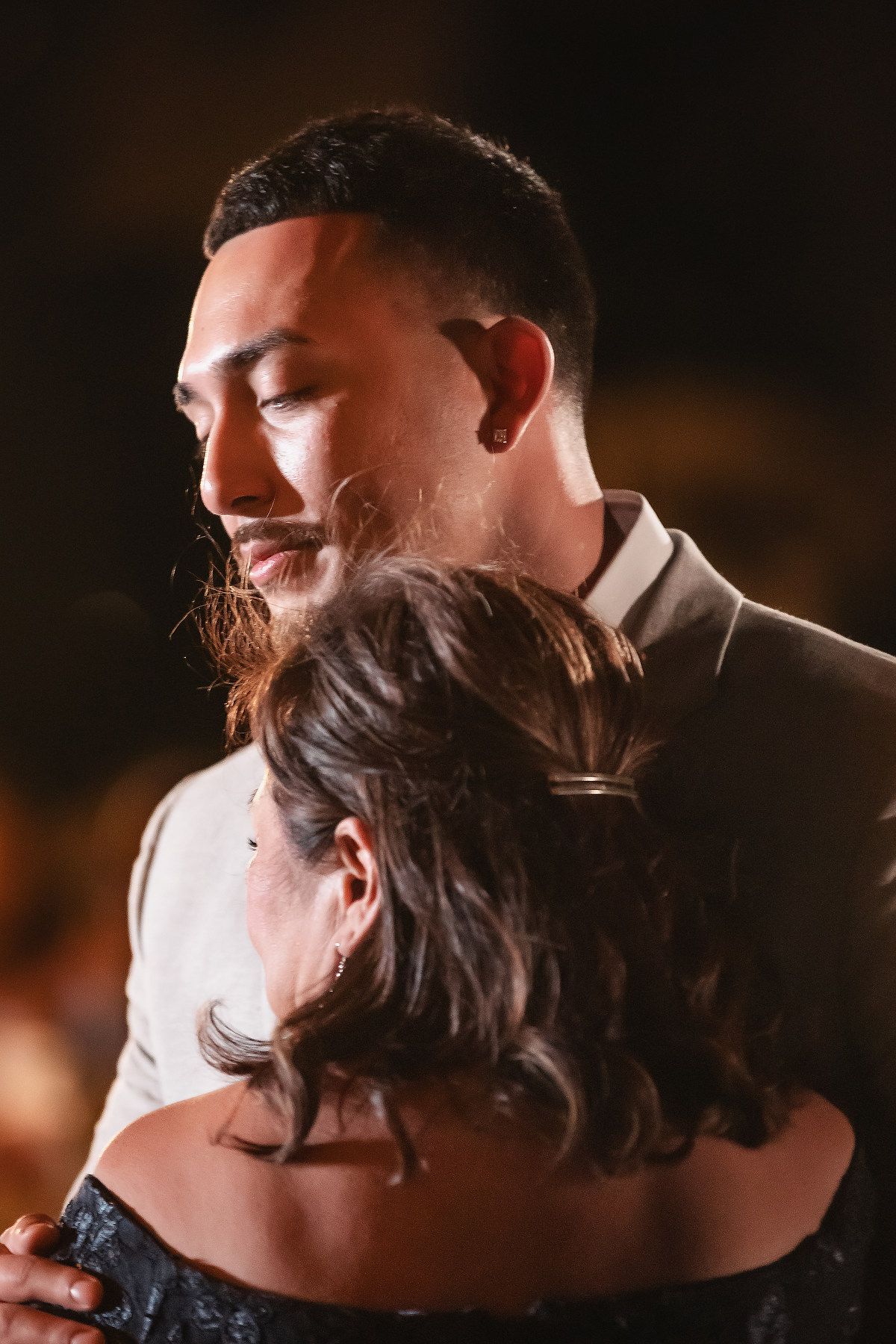 A groom with a beard embraces a woman in a tender slow dance moment at a dimly lit wedding reception.