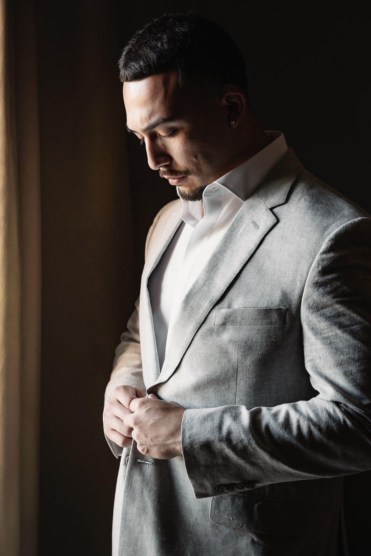 Groom in grey suit buttoning jacket near window in dramatic side lighting.