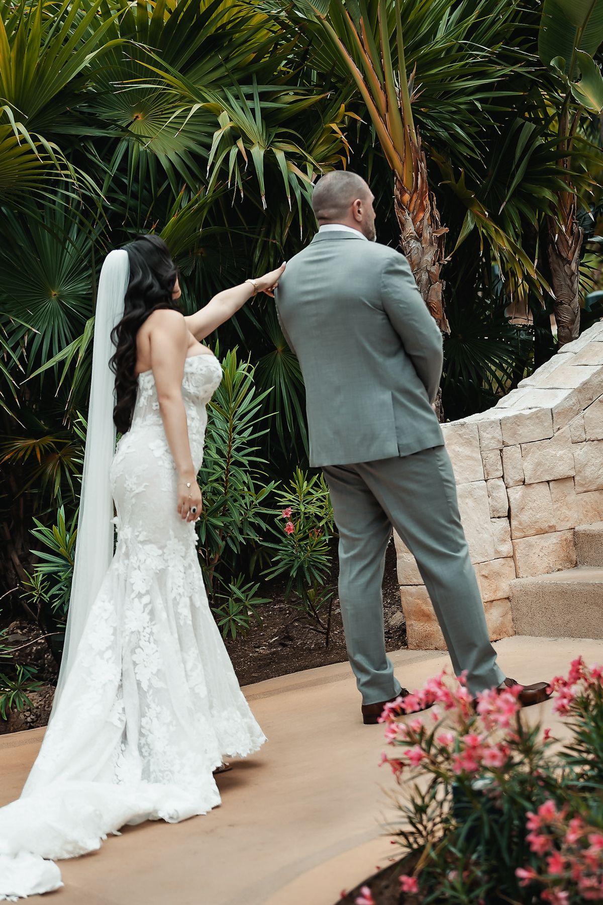 Bride in lace gown and veil taps groom in gray suit during first look moment surrounded by tropical garden.