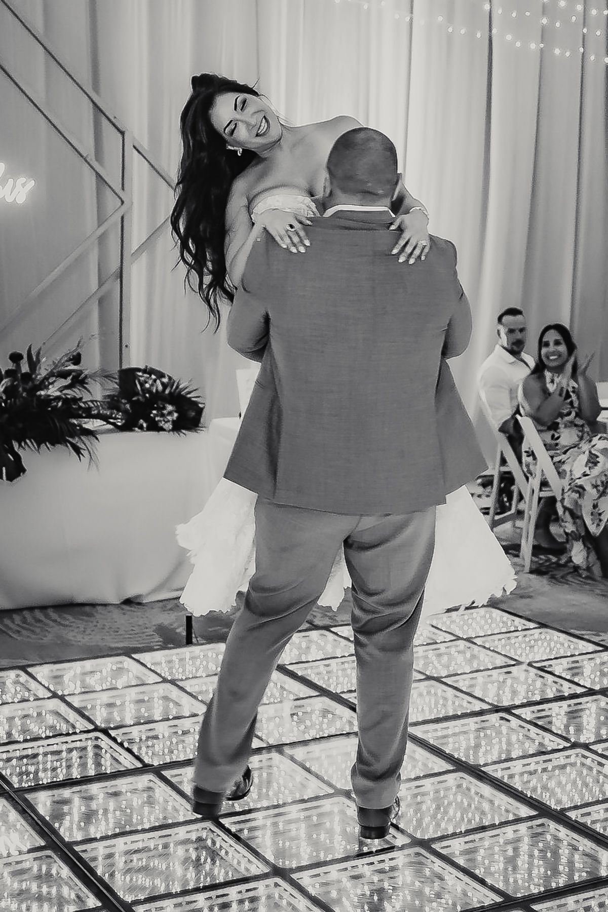 Groom lifts smiling bride during first dance at elegant wedding reception with mirrored dance floor.