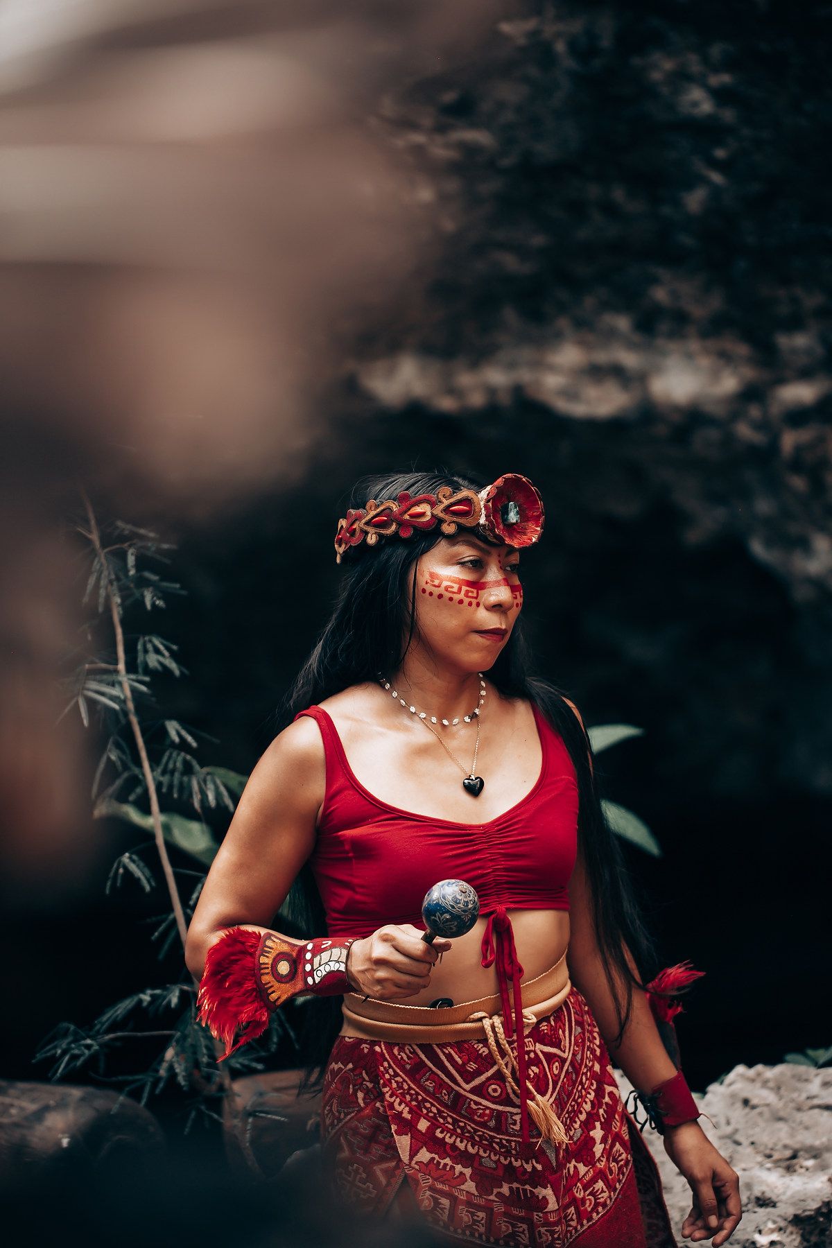 Woman in traditional indigenous red costume with face paint, headdress, and feather accessories holding a ritual object.