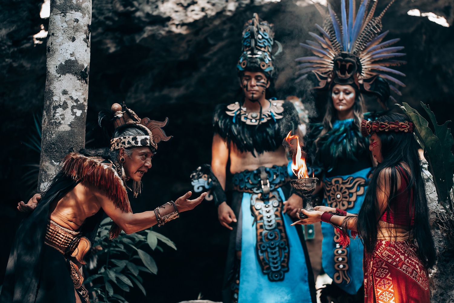 Four people in ornate Mayan ceremonial costumes perform a traditional ritual in a rocky cave environment.