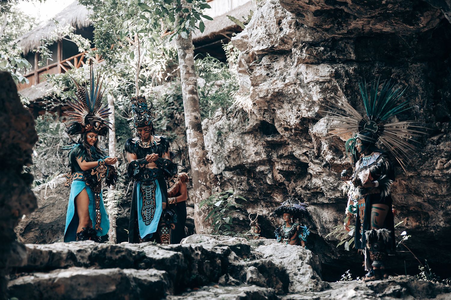 Three performers in traditional Mayan ceremonial costumes with feathered headdresses standing on rocks in a jungle setting.