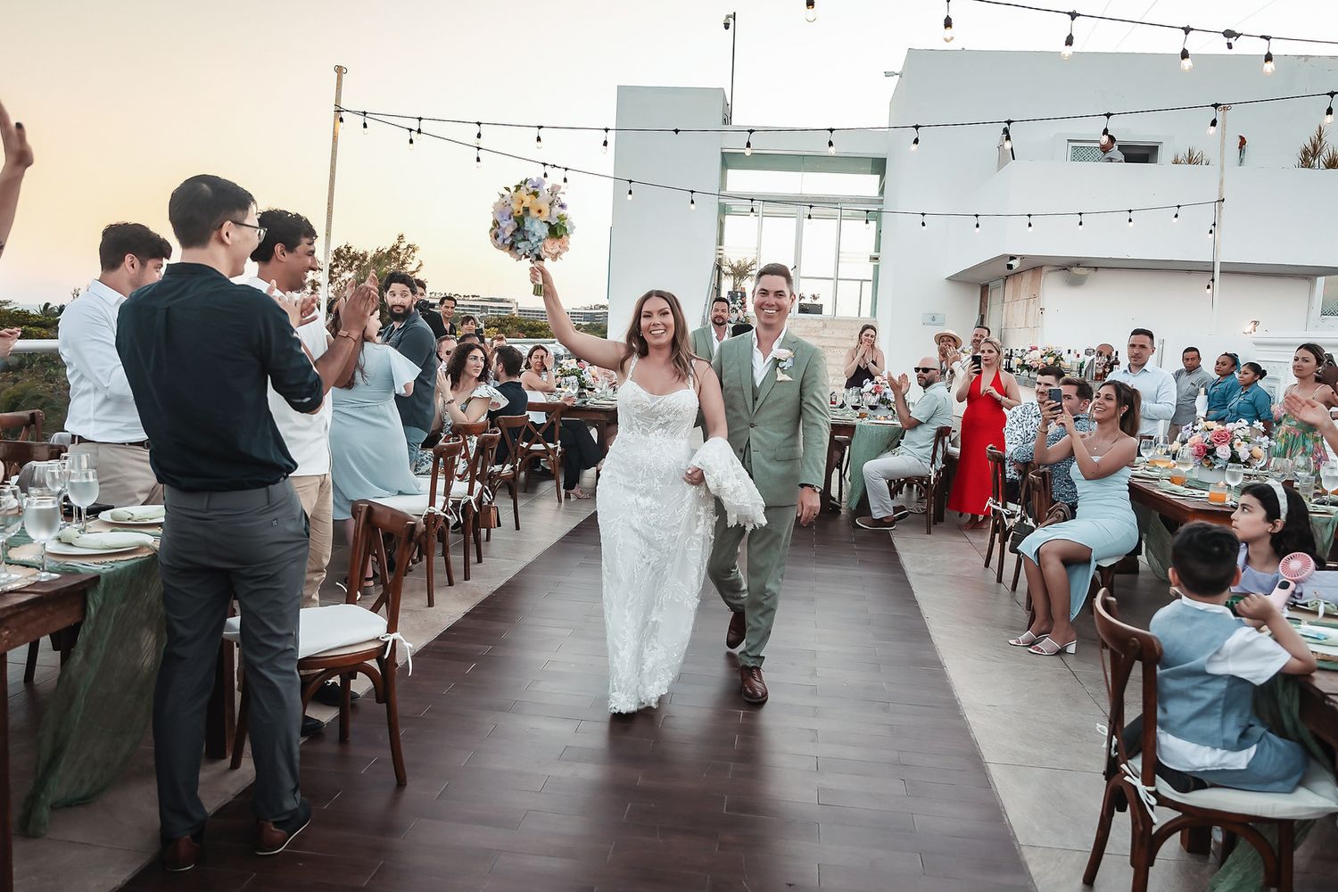 Bride and groom walk triumphantly down the aisle at an outdoor rooftop wedding reception with guests cheering.