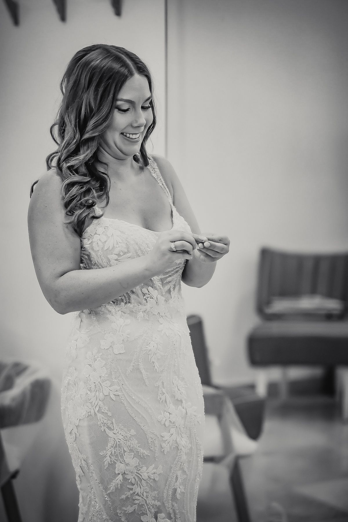 Bride in lace wedding gown smiling while adjusting dress details in black and white photo.