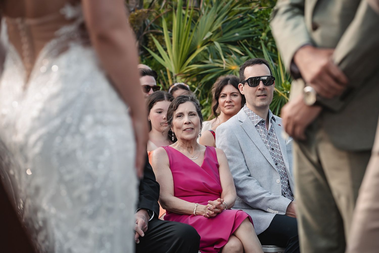 Wedding guests in formal attire watch an outdoor ceremony, with a woman in pink dress seated prominently among the audience.