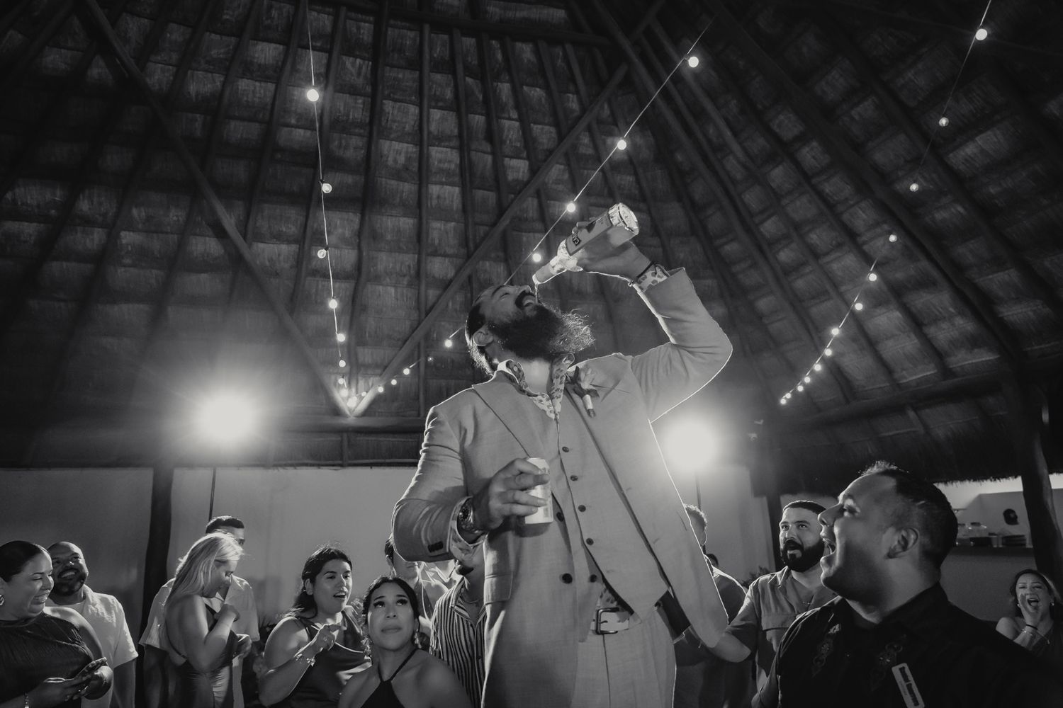 Groom celebrates joyfully at wedding reception in black and white photo with string lights and guests.