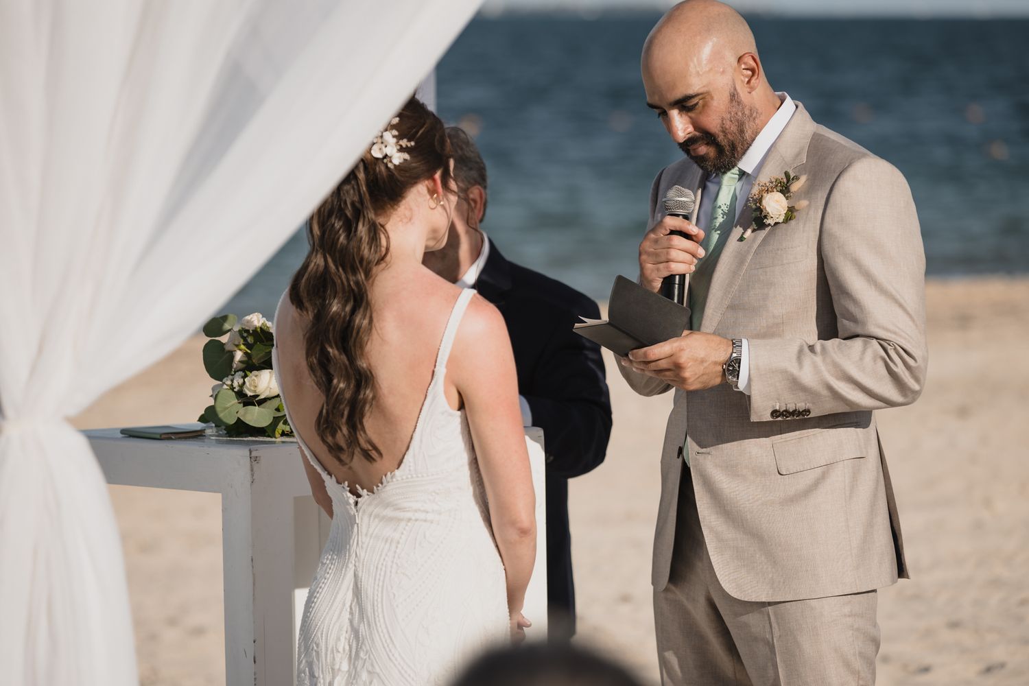 Groom reading vows to bride at elegant beach wedding ceremony with officiant.