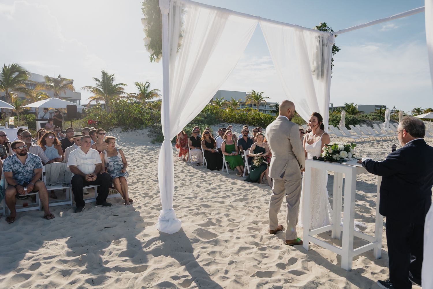 Outdoor beach wedding ceremony with guests seated on sand beneath white draped arch canopy in tropical setting.