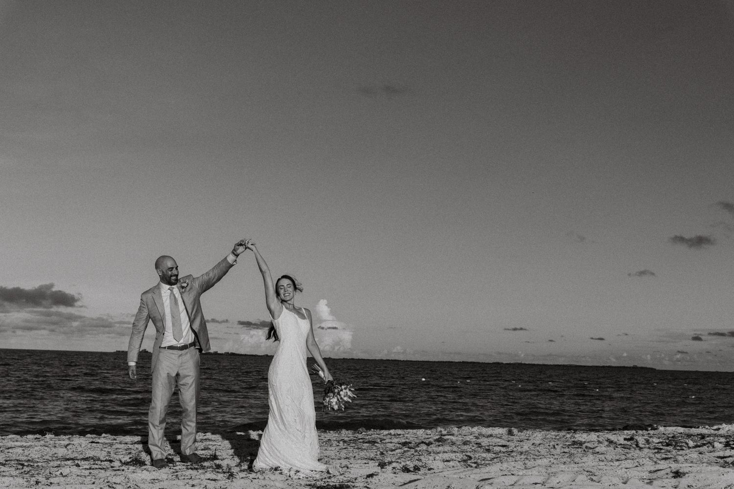 Black and white beach wedding photo of bride and groom dancing joyfully by the ocean shore.
