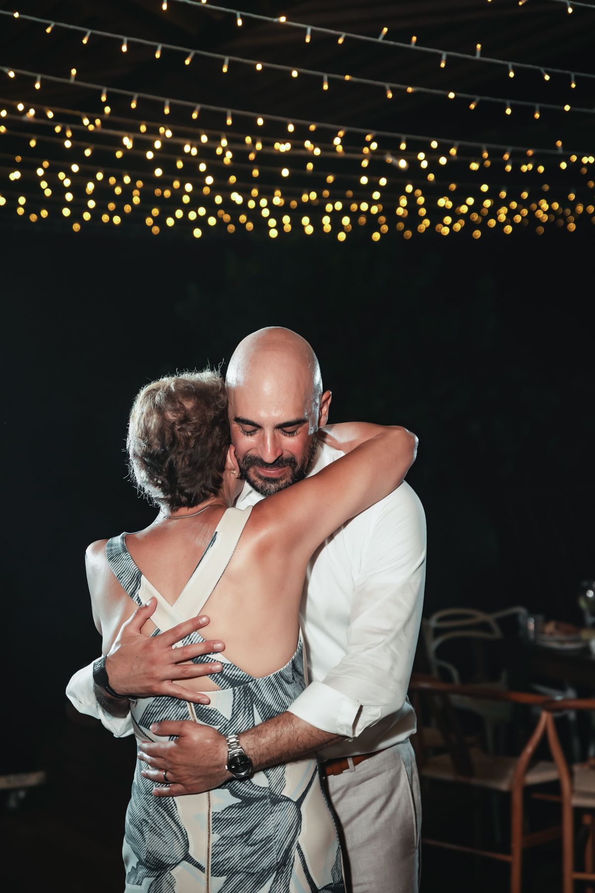 Bald groom in white suit shares emotional dance with woman under warm string lights at outdoor wedding reception.