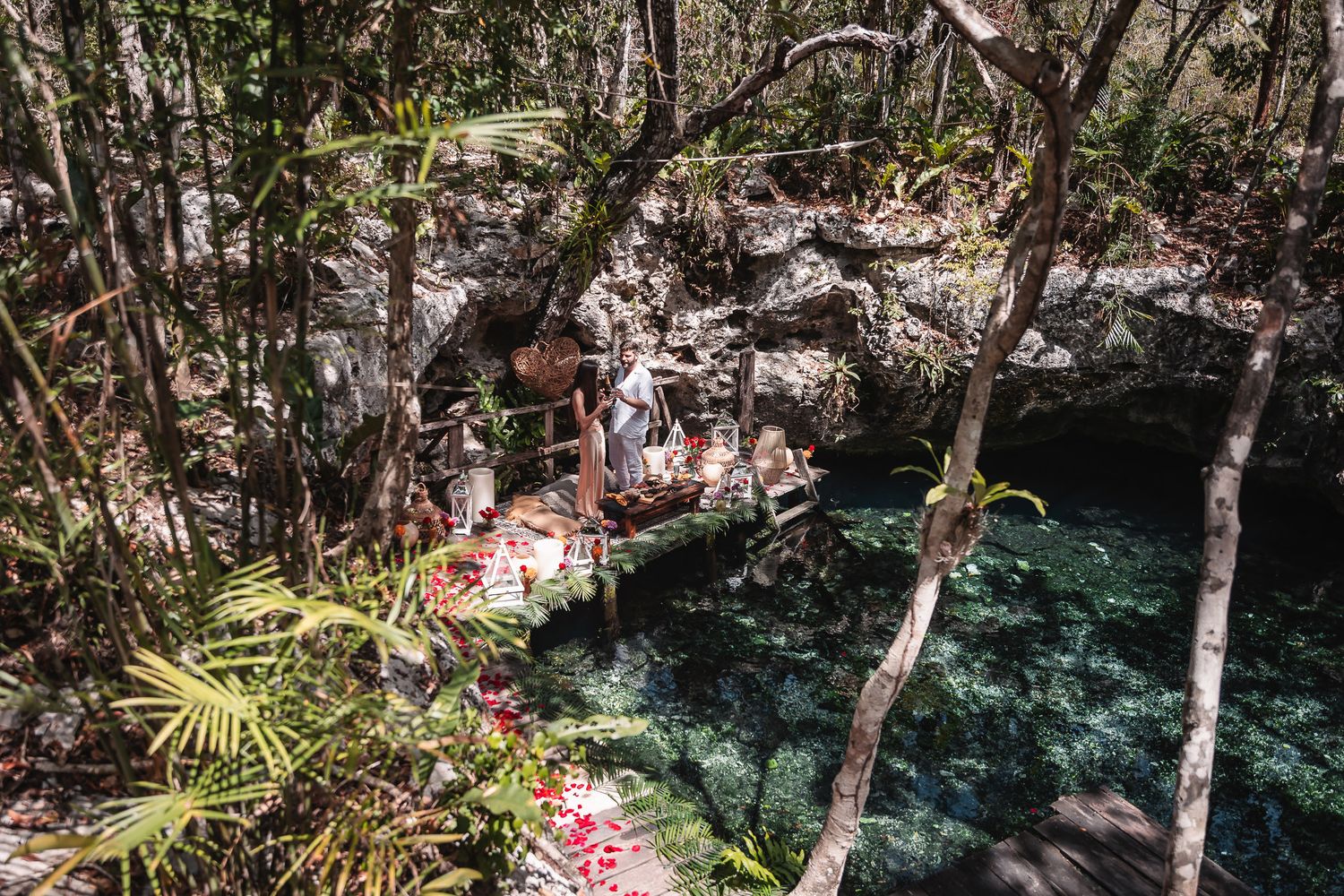 Lush jungle cenote with crystal-clear turquoise water, surrounded by tropical trees, rocks, and red flower petals near Tulum, Mexico.