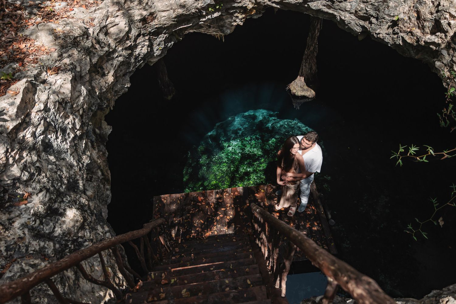 Couple standing at the entrance of a cave with wooden stairs leading down to a lush cenote.