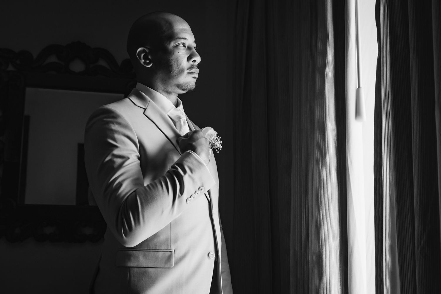 Black and white portrait of groom in suit adjusting tie near window light.