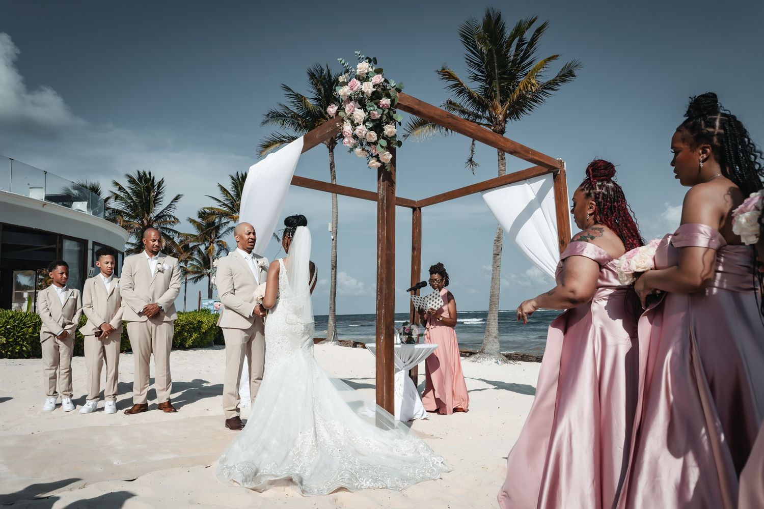 Beach wedding ceremony with bride and groom at floral arch, groomsmen and bridesmaids in pink dresses, ocean backdrop.
