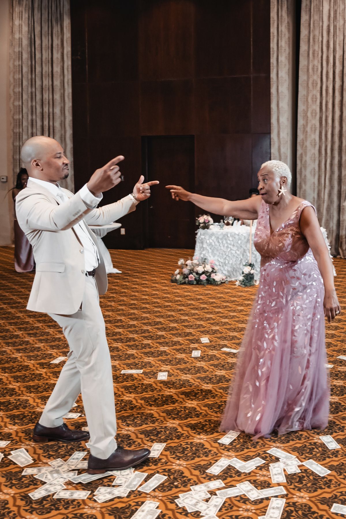 Man and elderly woman in pink gown point at each other playfully on wedding reception dance floor.