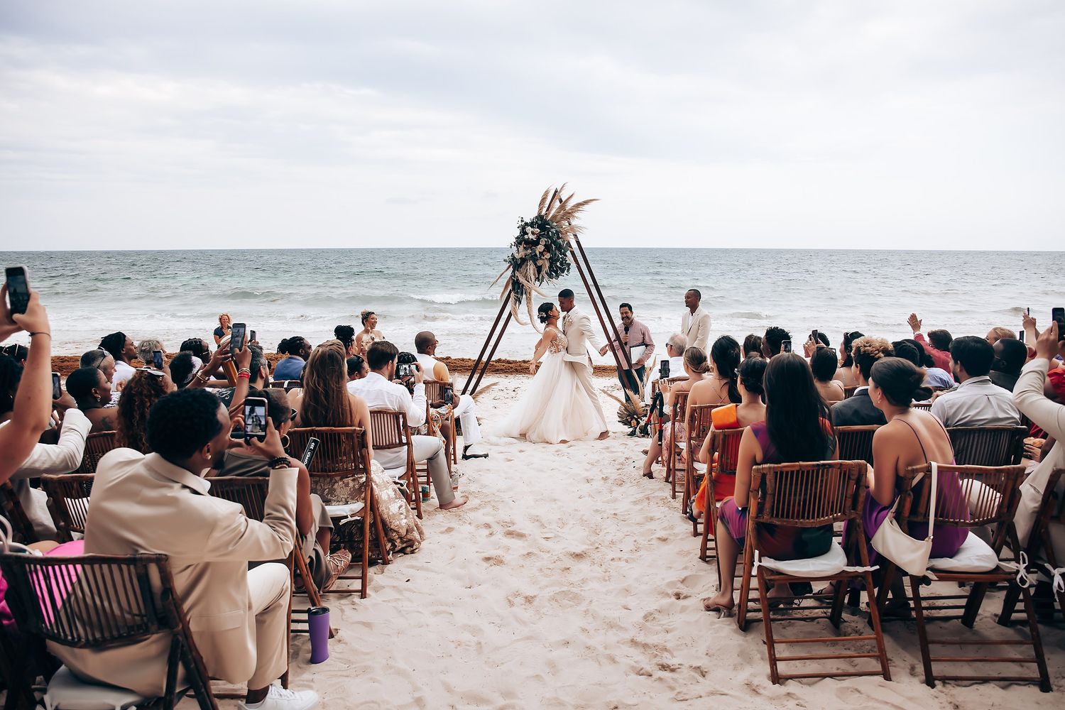 A beachfront wedding ceremony with wooden arch and guests seated on both sides.