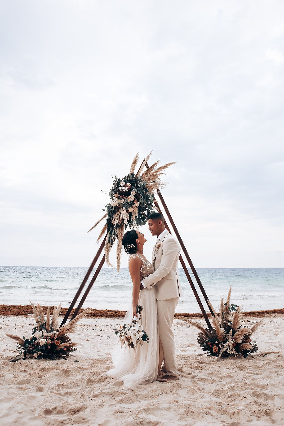 A beach wedding portrait under a bohemian triangle arch decorated with dried flowers.