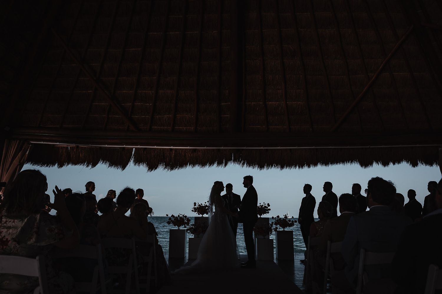 Silhouettes of wedding guests dancing under a dimly lit thatched roof.