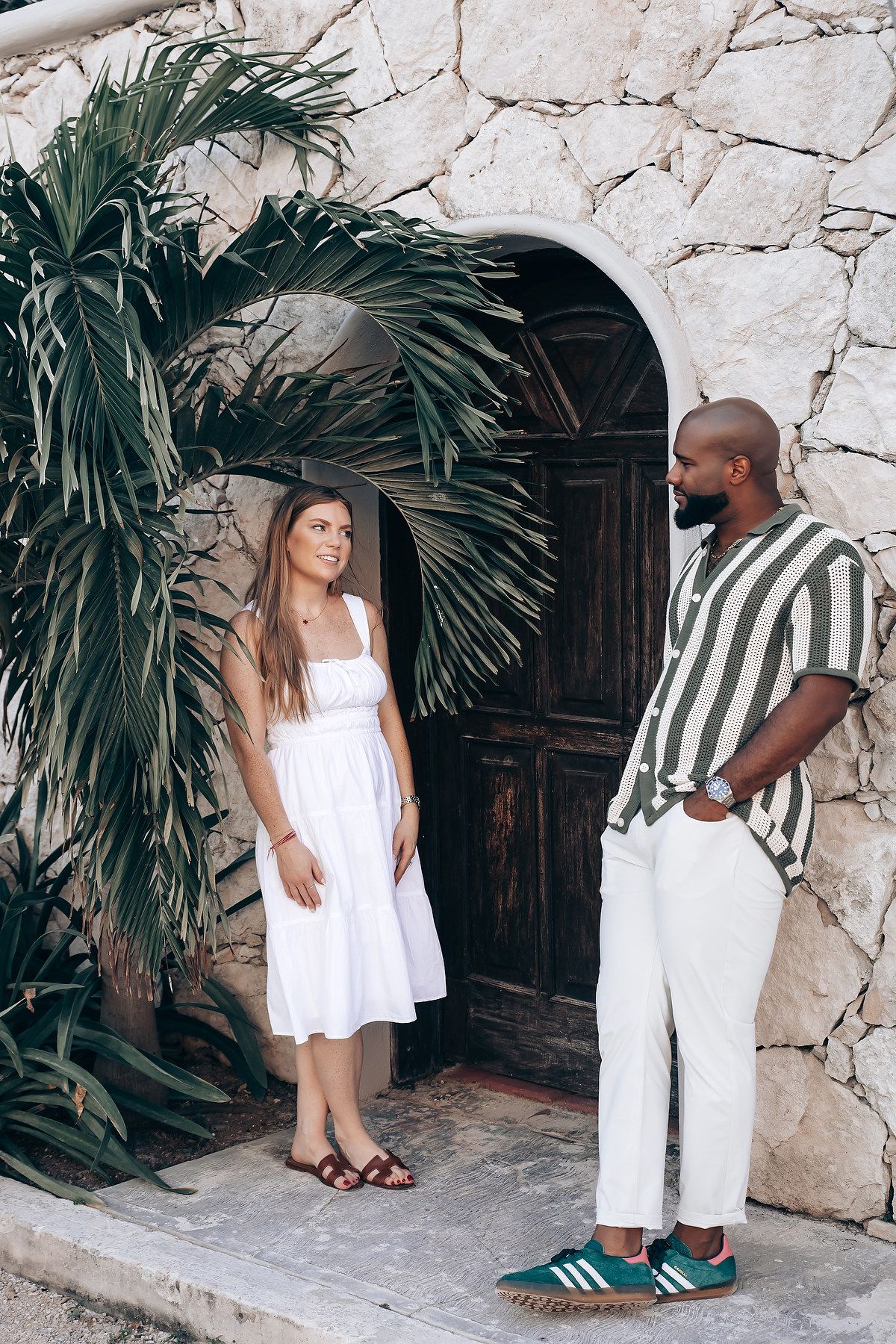 Couple standing by a rustic stone archway with wooden door, woman in white dress, man in striped shirt and white pants.