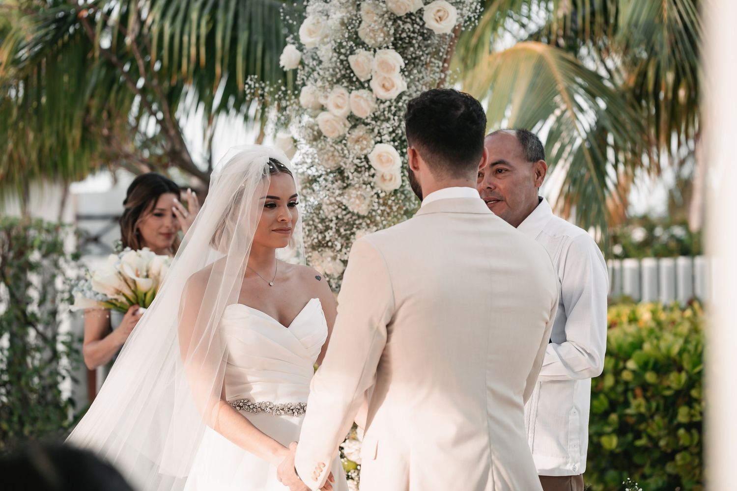 Bride and groom exchanging vows at outdoor wedding ceremony beneath white floral arch with palm trees.