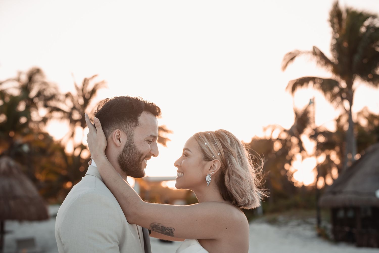 Bride and groom share a romantic moment at sunset on a tropical beach with palm trees.