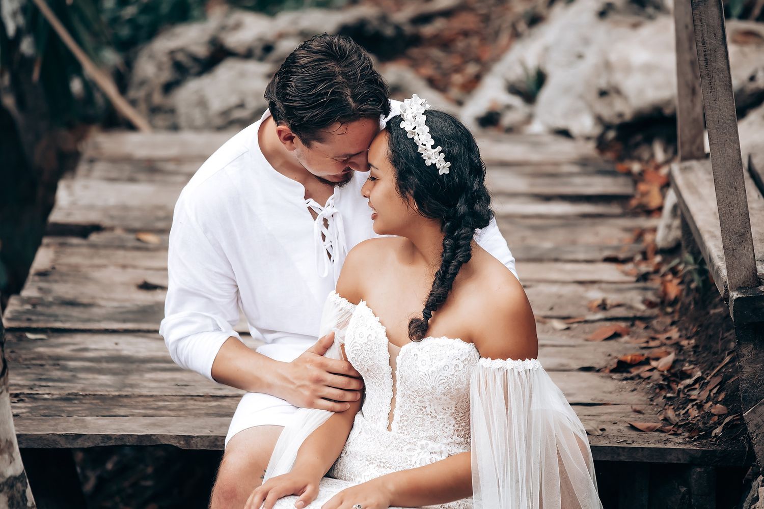 A romantic couple in white attire share an intimate moment on a rustic wooden walkway surrounded by trees.