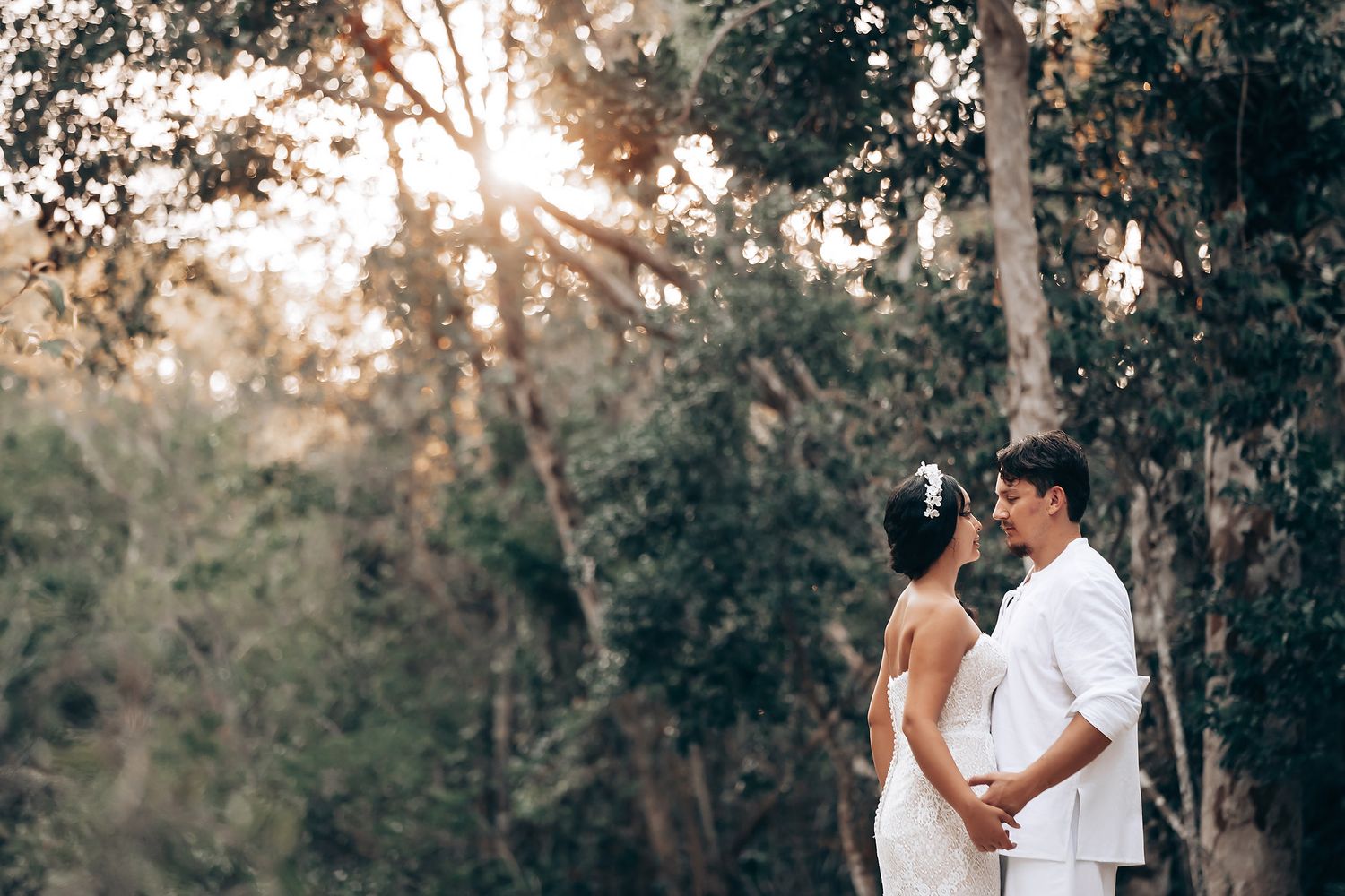 A couple in white outfits embraces during a romantic outdoor photoshoot among eucalyptus trees at sunset.