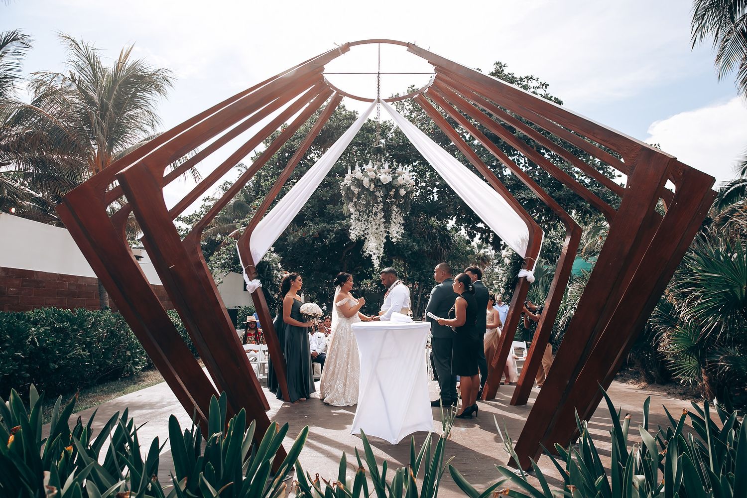 Wooden arch decorated with flowers stands against blue sky at tropical beach wedding venue.