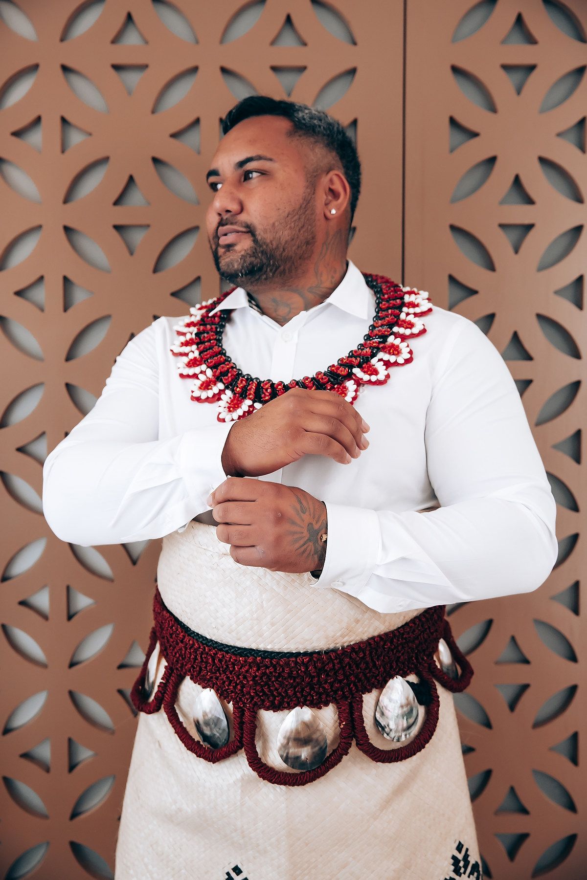 A person in a white outfit wearing a traditional red beaded necklace poses against a geometric patterned wall.
