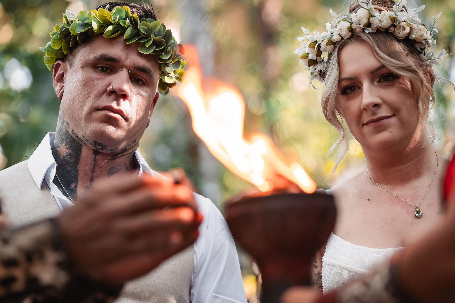 Couple in floral crowns holding flames during an outdoor wedding ceremony.