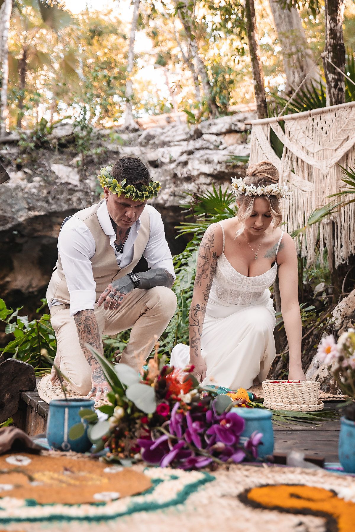 Tattooed bride and groom in floral crowns kneel during outdoor bohemian wedding ceremony in tropical forest setting.