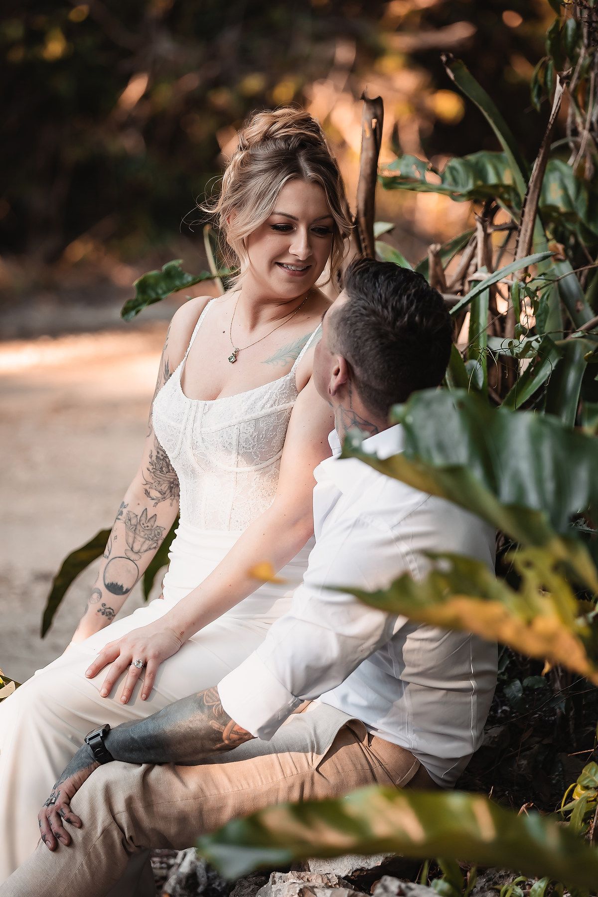 Tattooed bride in white dress smiling at groom while sitting among tropical plants in golden light.