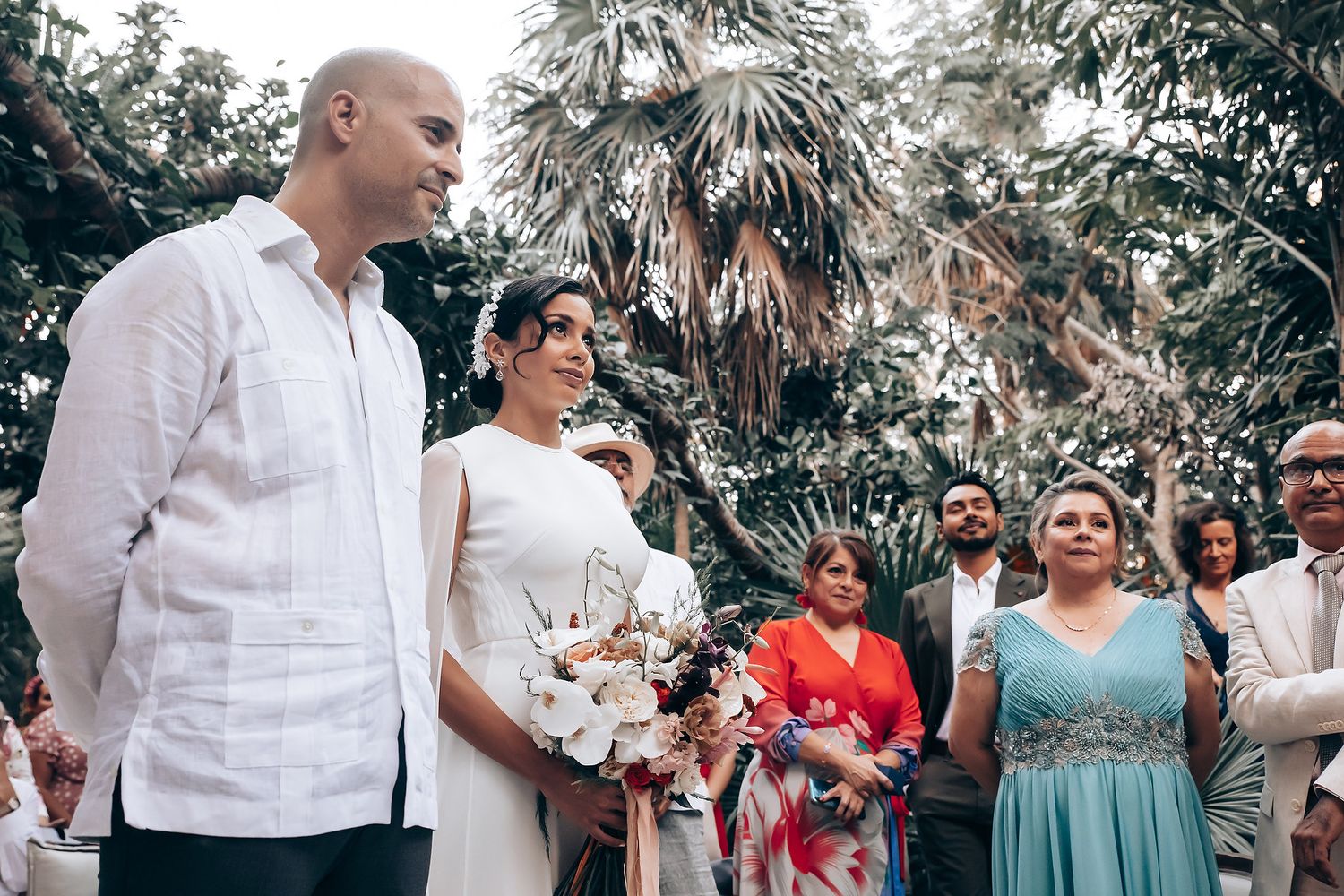 Wedding party gathered outdoors under palm trees for group photos.