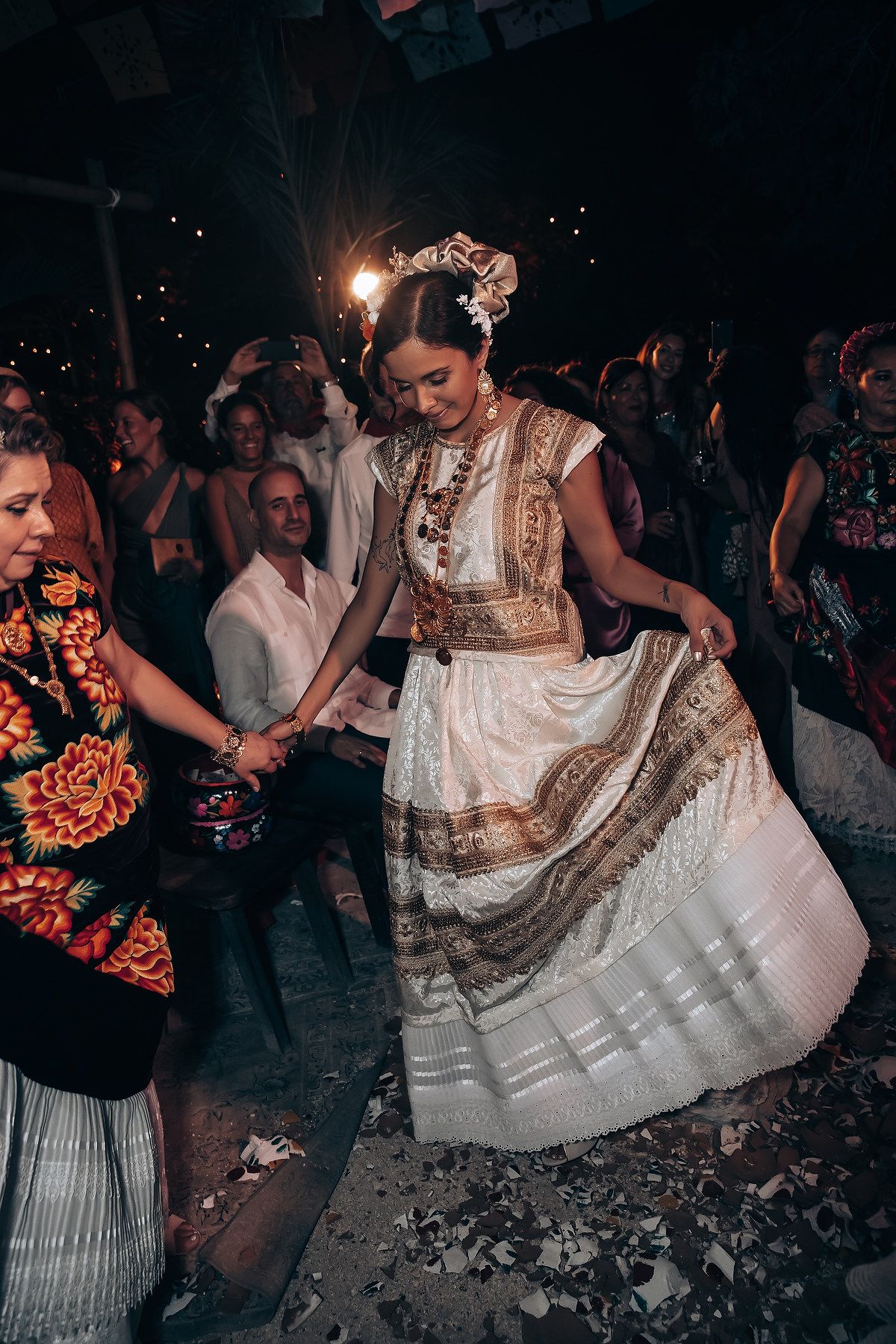 Traditional dancers in white dresses performing at a nighttime celebration with dramatic lighting.