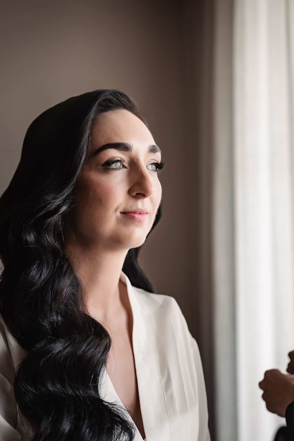 Bride with dark wavy hair gazes toward window light wearing white robe before wedding ceremony.