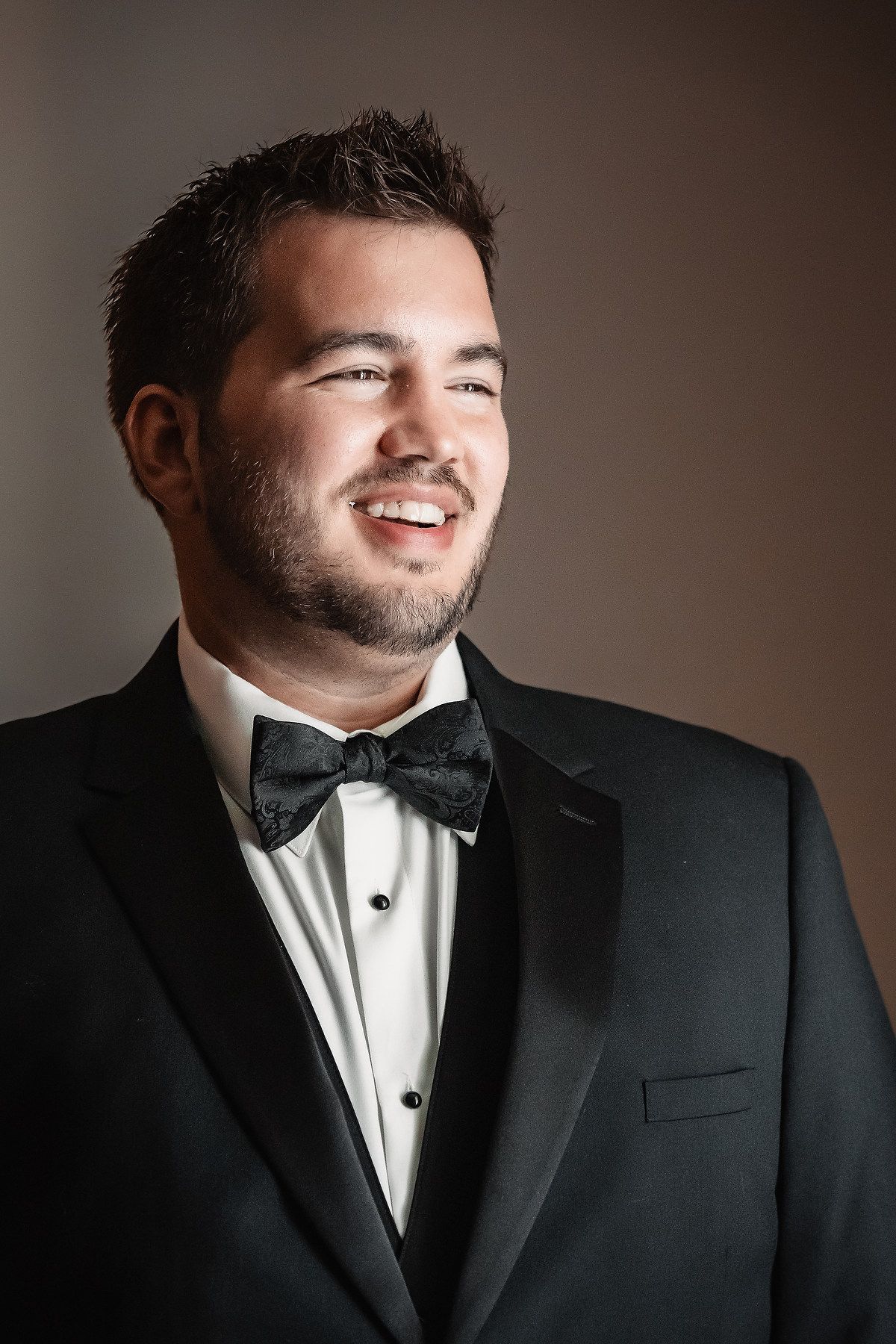 Smiling groom in black tuxedo with bow tie posing for formal wedding portrait.