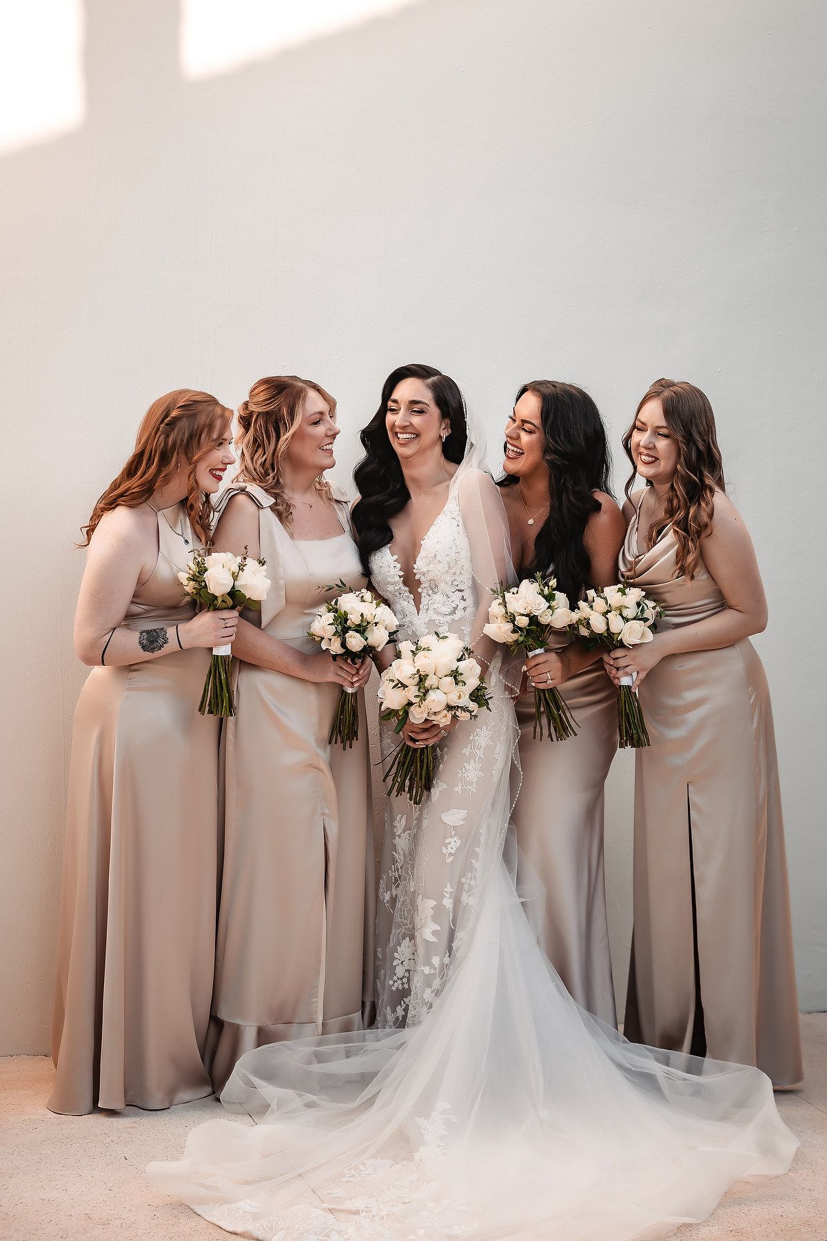 Bride in lace gown with four bridesmaids in champagne satin dresses holding white bouquets, smiling together.