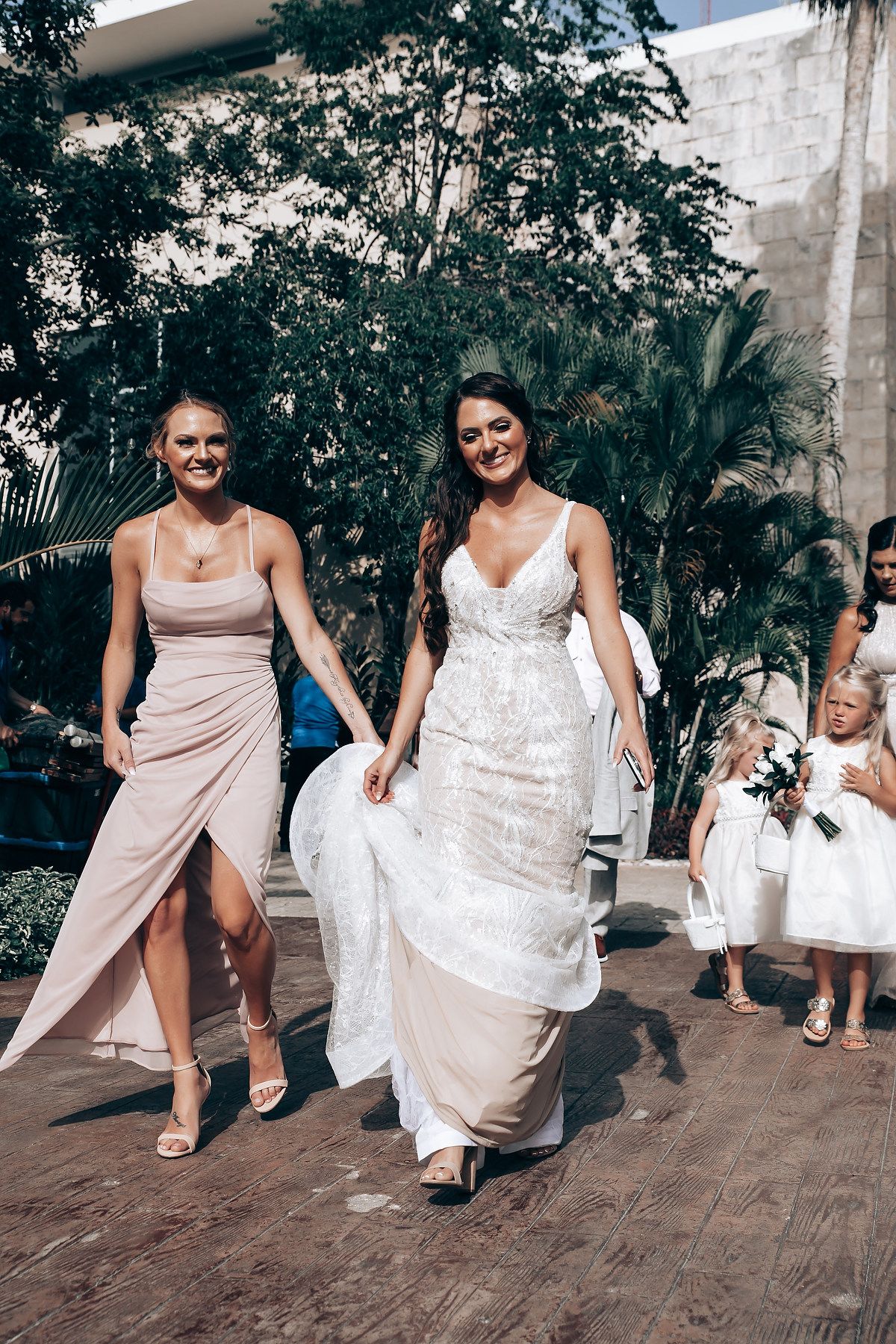 Wedding party walking outdoors with bridesmaids in blush gowns and flower girls in white dresses.