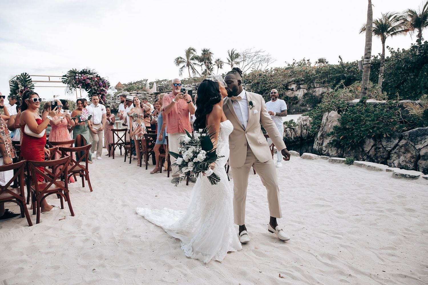 Beach wedding ceremony with guests seated in white chairs along sandy shoreline.